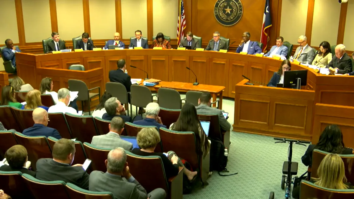 Mike Morath sits on a table and speaks into a microphone in front of Texas House lawmakers