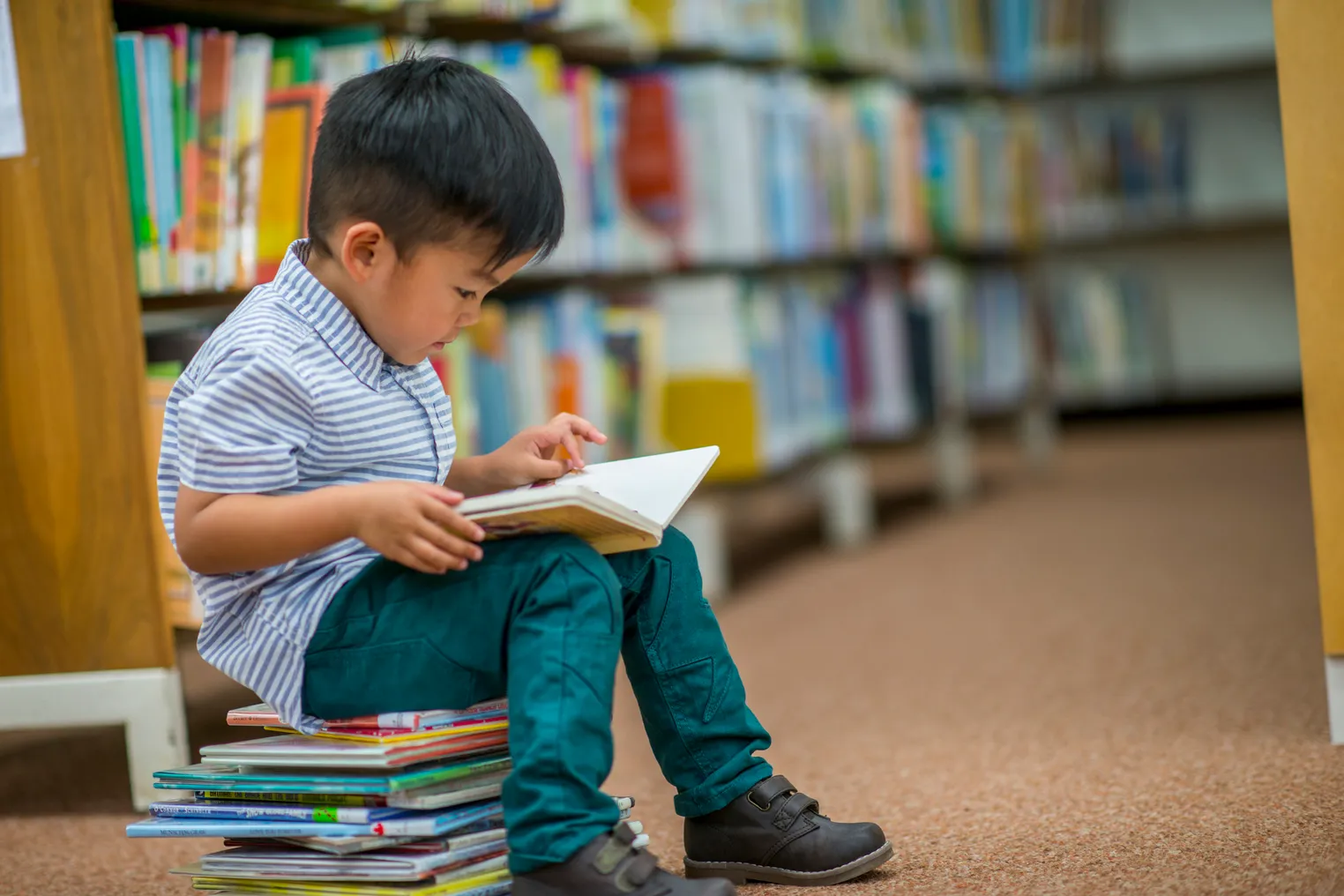 A young child sits on a stack of books near shelves of books. Child has a book on lap and is looking at book.