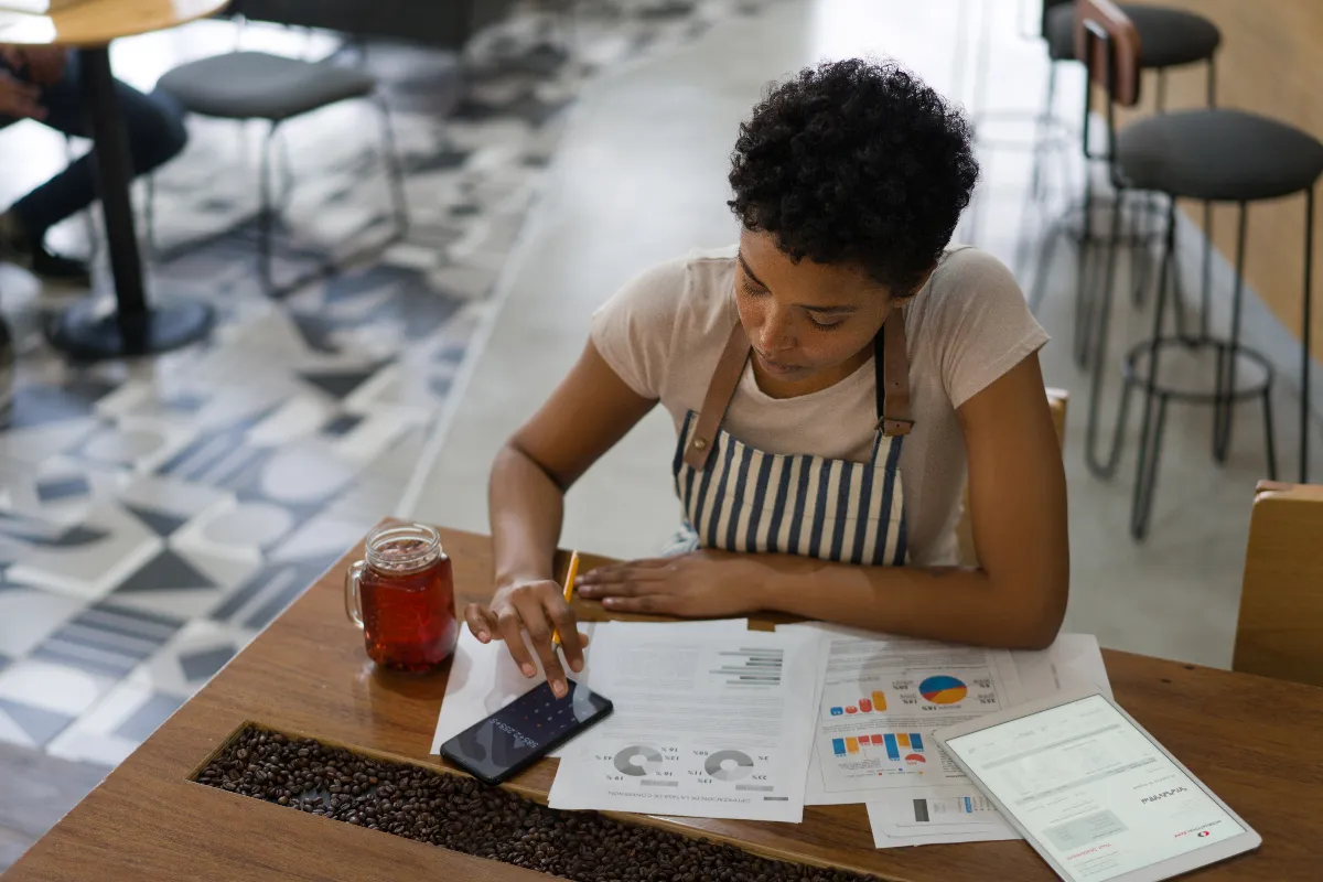 A woman sitting at a table using her phone as a calculator. On the table there are financial charts.