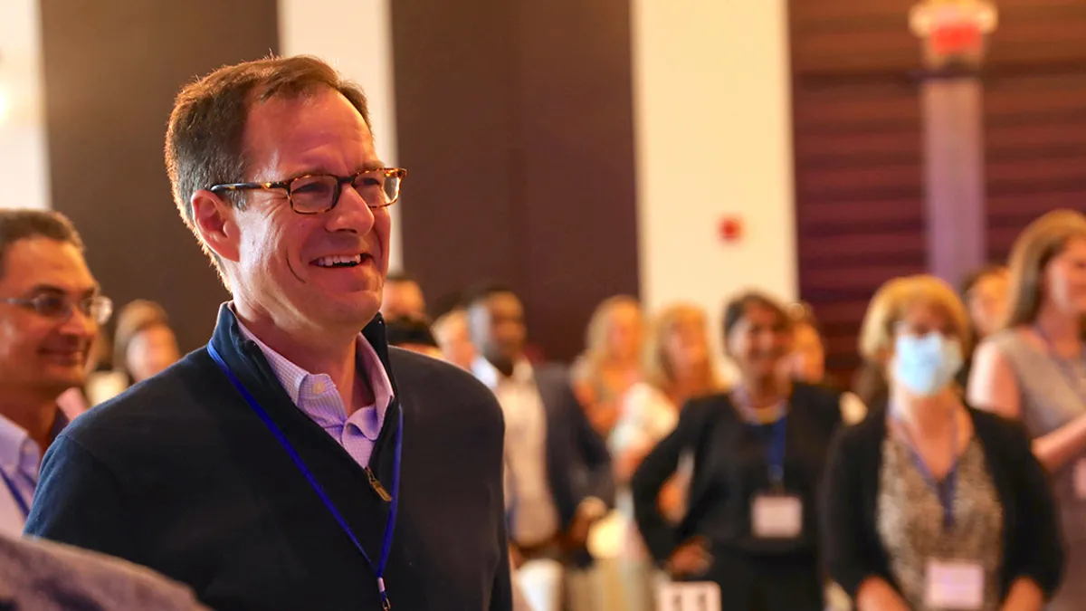 Brian Goff stands laughing in the forefront, with a crowded room of business attire-clad people in the background.