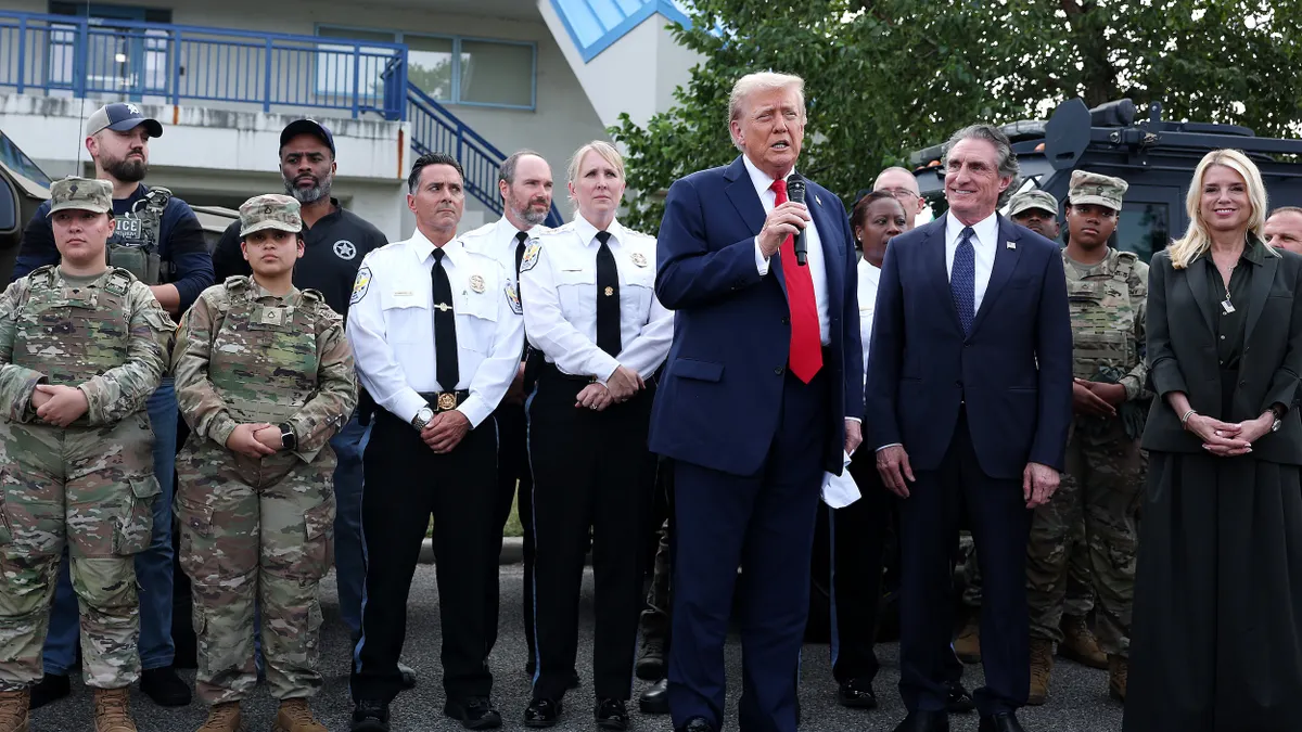 President Trump speaks into a microphone in front of people wearing military and police uniforms, a man in a suit and a woman dressed in black.