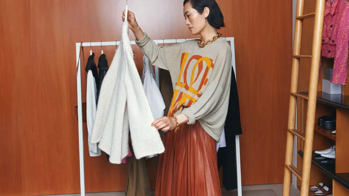 A person holds a sweater on a hanger in front of a rack of other clothes.