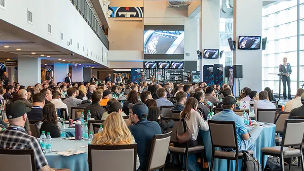 A shot of a group of people sitting at circular banquet tables amid a crowd. A podium sits to the right of the image.