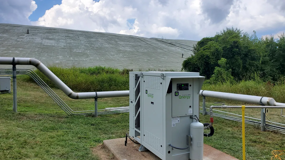 A small metal box with a tank sits in front of a pipeline, with a landfill in the background.