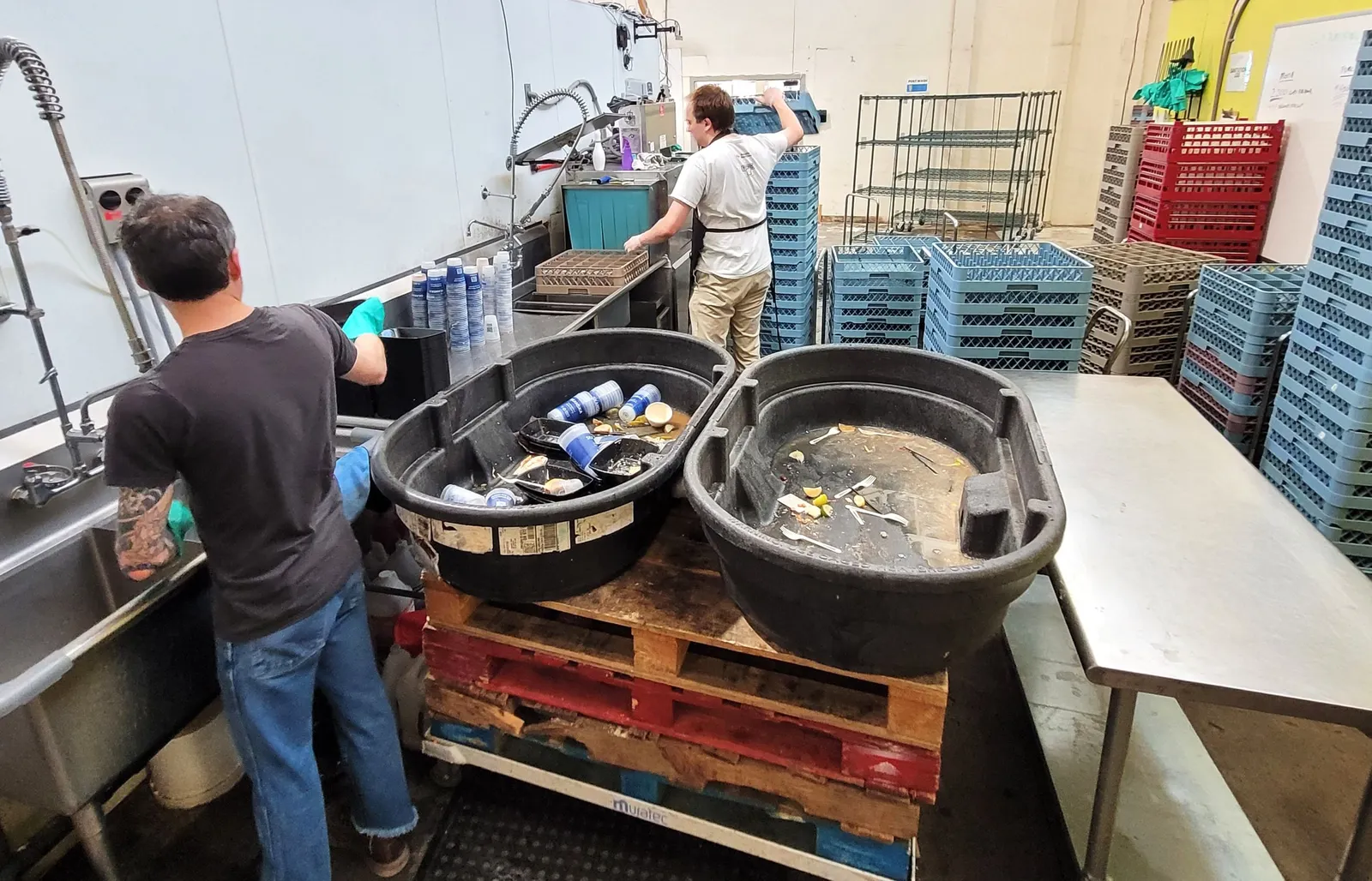 Two people work at a commercial washing station near stacked foodservice ware.