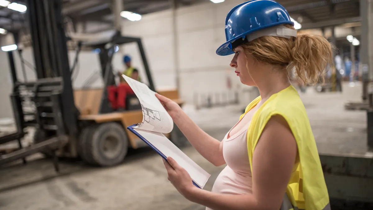 A pregnant person in a hard hat and construction vest reads a clipboard