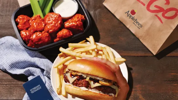 A photograph of a hand holding up a cheeseburger above a white plate. The plate is on a wooden table and there is a blue card on the left that says Wyndham Rewards. On the right there is a paper bag that says Applebee's To-Go.