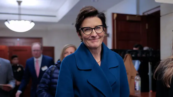 Jane Fraser, wearing a blue coat, walks down a hallway with people surrounding her, including a person holding a mobile phone in front of her.