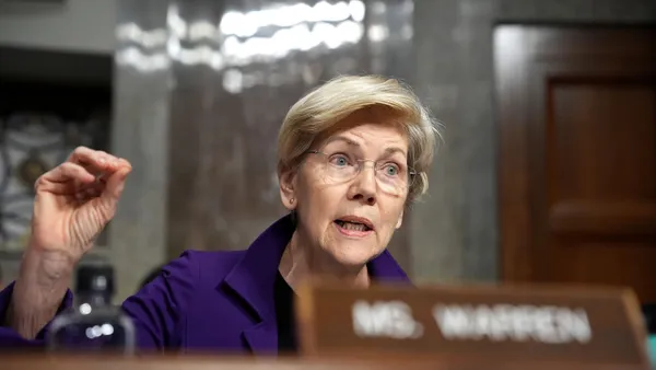 Senator Elizabeth Warren speaks during a legislative hearing, holding her handout to the side. A sign in front of her reads "Ms. Warren."