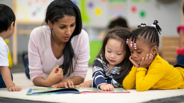 An adult sits at a table in a classroom looking at a book on the table. Young students are sitting near the adult.