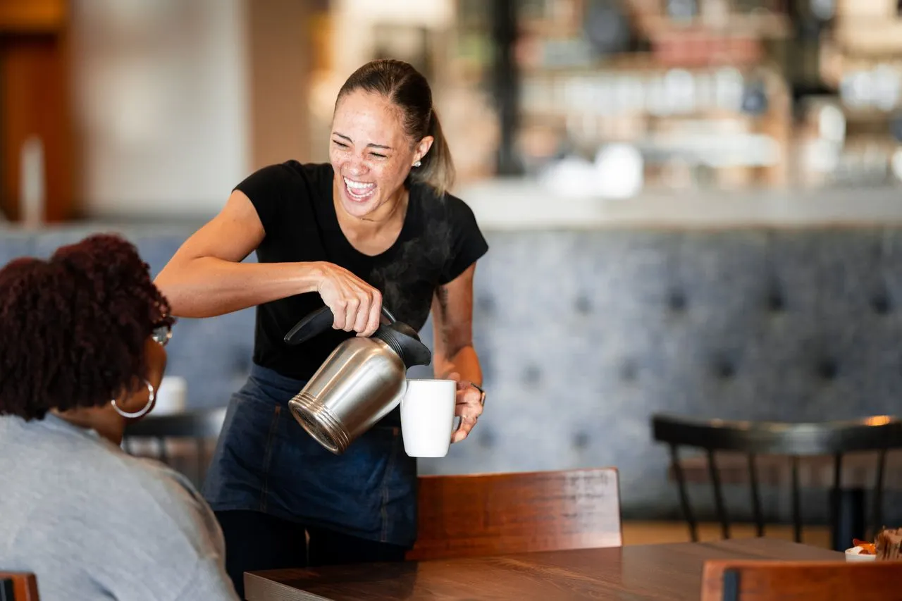 A person wearing black pours coffee into a white mug inside a restaurant. There is another person sitting at the table with her back to the camera.