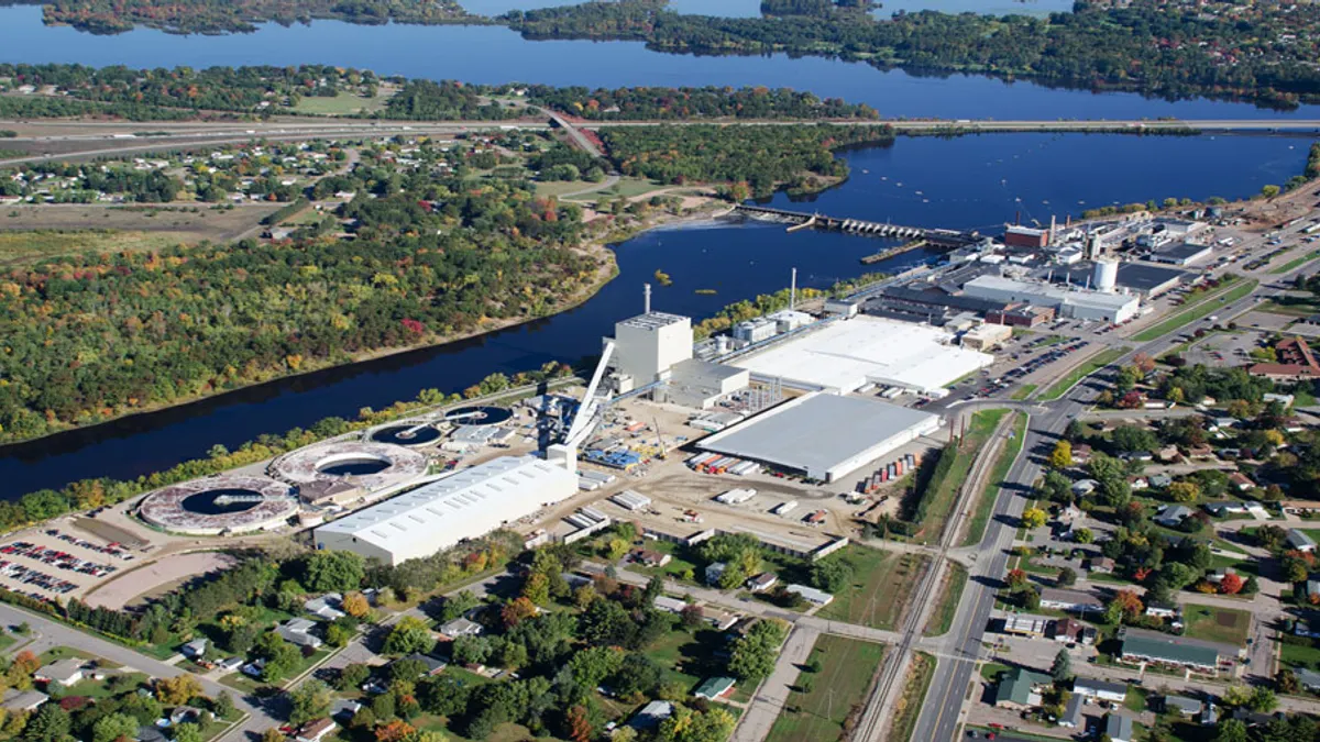 An aerial photograph shows a river flowing by a sprawling mill with a lake behind a dam in the background.