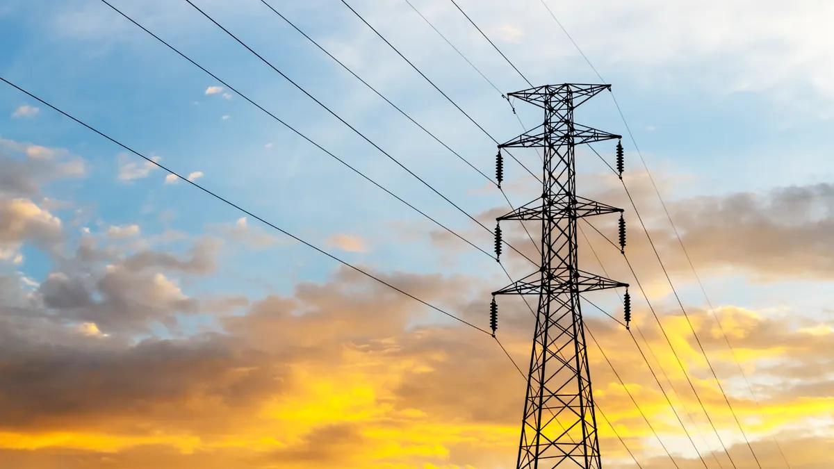 Silhouette of a large electrical transmission tower with power lines against a dramatic sunset sky featuring blue and bright orange clouds.
