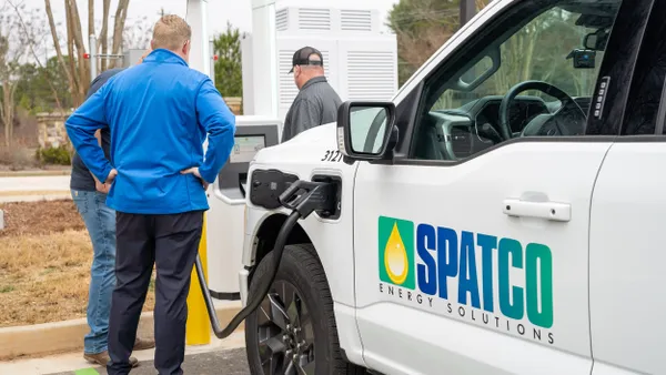 A photo of two people looking at an outdoor electric vehicle charging cabinet, with one of the cabinet's connectors hooked into the charging port on a truck. Lettering on the truck reads "SPATCO."