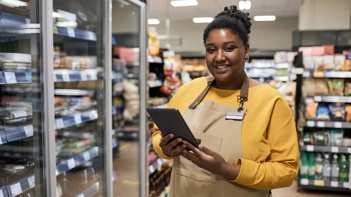 Person holding a tablet standing next to a glass refrigerator in a convenient store.