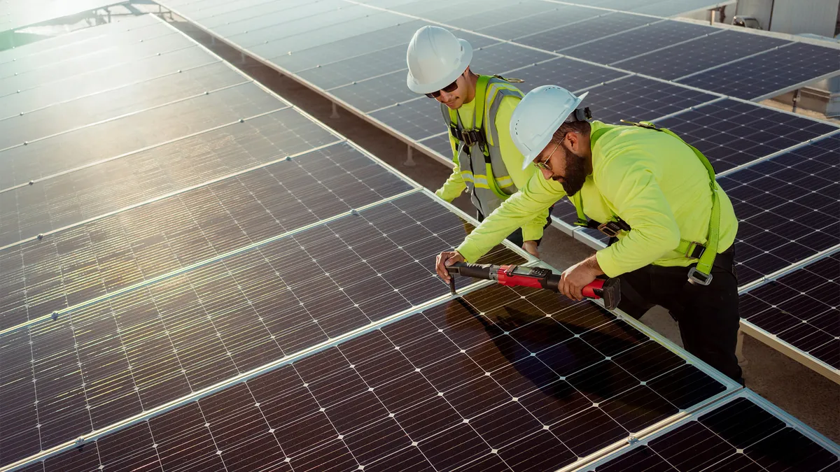 Two men in yellow vests and hardhats install a solar panel.