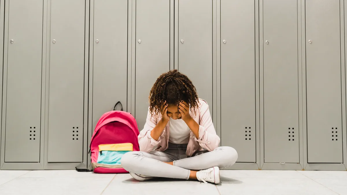 Young student sits in front of lockers inside a school building with head in hands.