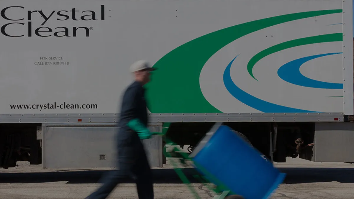 Person in a uniform pushing a large, blue drum on a dolly, in front of a tractor trailer.