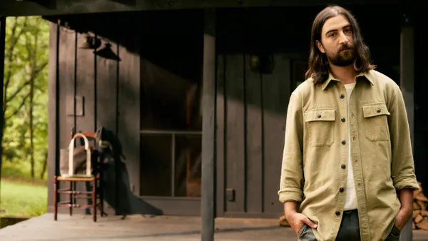 Musician Noah Kahan stands in front of a shed. He is wearing a button down shirt from LL Bean, and on the porch of the shed other LL Bean products are placed.