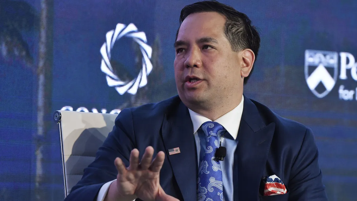 Utah State Attorney General Sean Reyes sitting and speaking with his right hand raised, an american flag pin on his right lapel, a flag handkerchief in right suit breast pocket and blue paisley tie