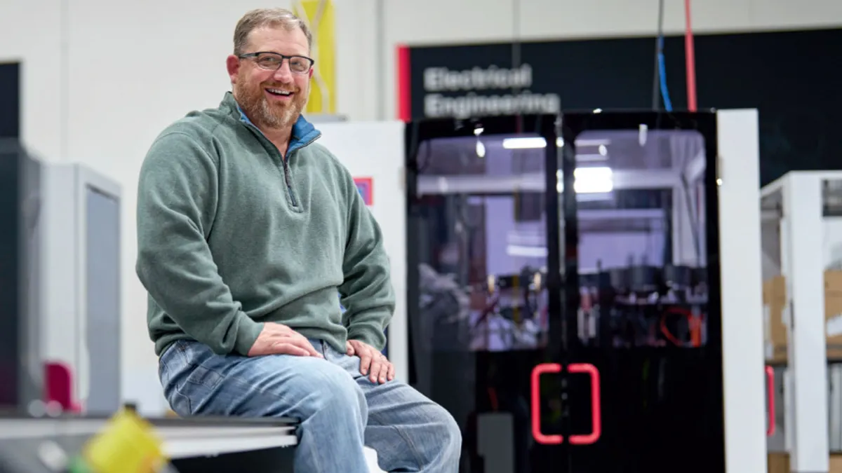 A smiling man wearing glasses and a green sweater sits in an engineering workshop beside equipment labeled “Electrical Engineering.”
