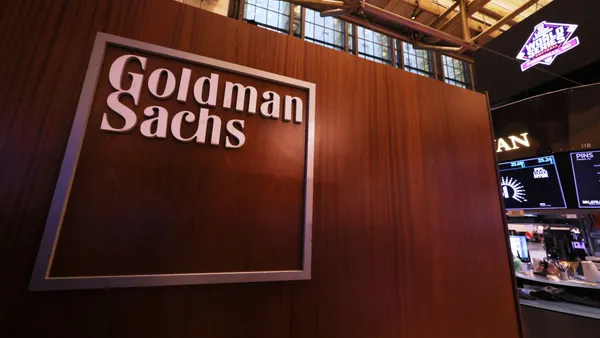 The words "Goldman Sachs" are seen on a partial wall with a wood finish.