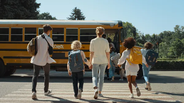 Students with backpacks and an adult are walking toward the side of a yellow school bus. The bus doors are open and students are going inside the bus.