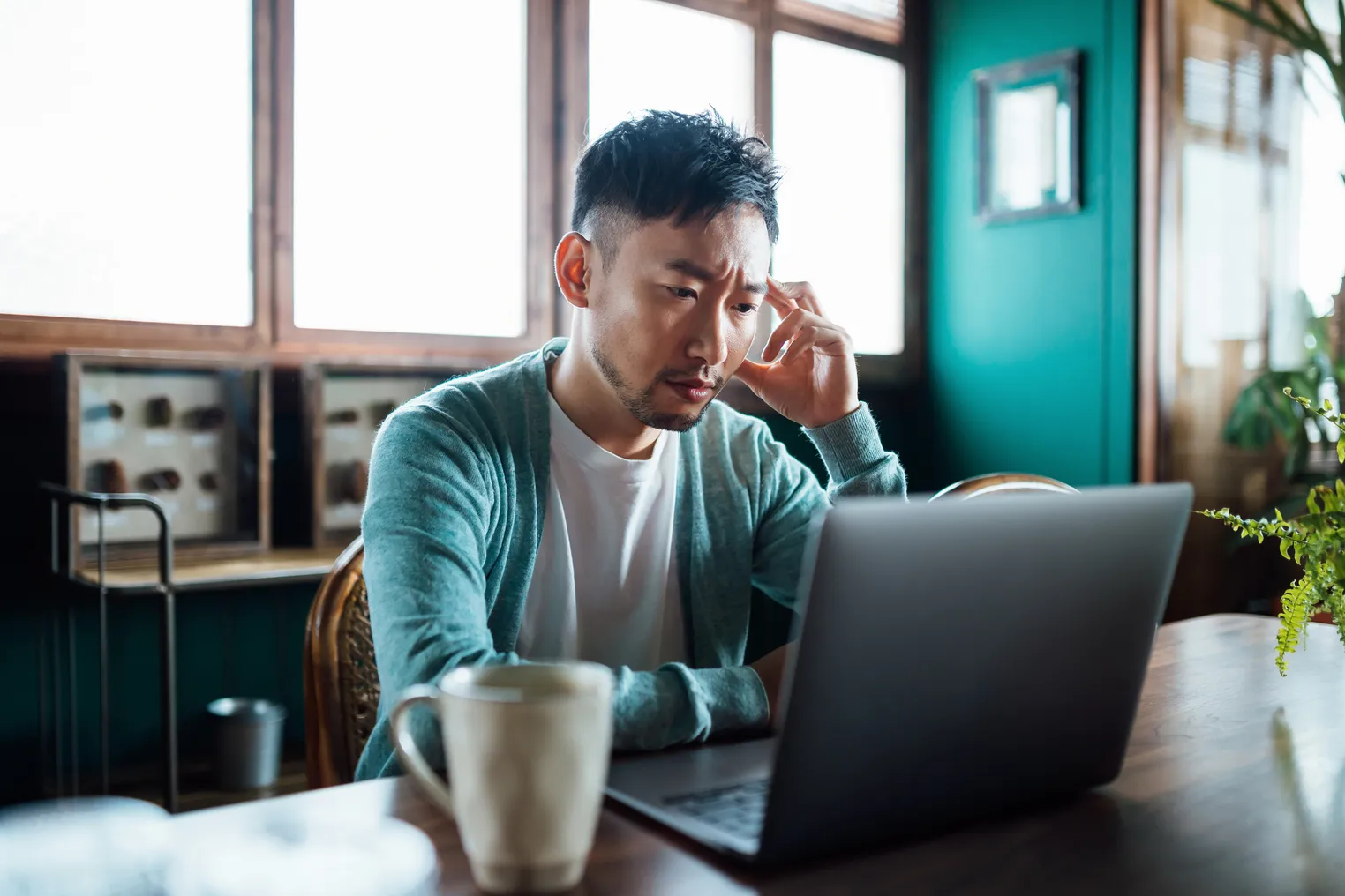 Worried Asian man with his hand on head, using laptop computer at home, looking concerned and stressed out