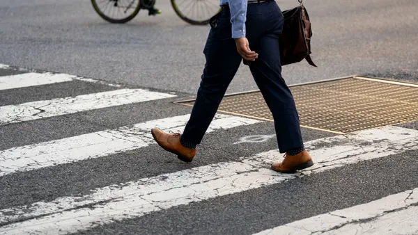 A person wearing slacks and dress shoes and carrying a shoulder bag moves through a crosswalk.