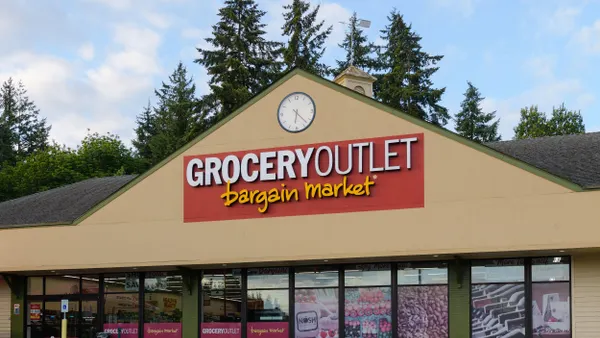 A tan gable-roofed building with a clock, red and yellow “GROCERY OUTLET bargain market” sign, and large window graphics showing produce.