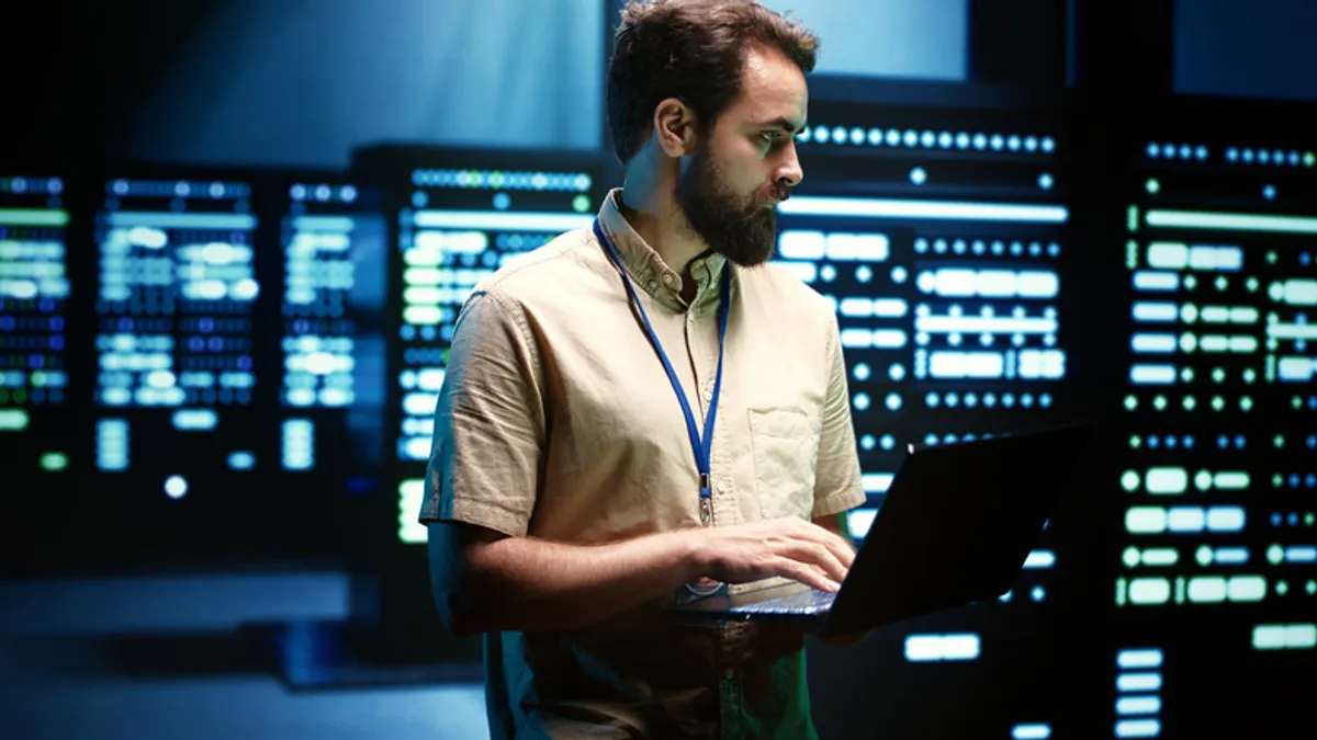 Man with beard wearing a tan shirt and blue lanyard works on a laptop in front of glowing server racks.