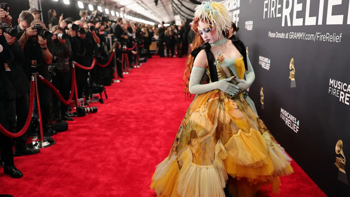 A person stands on a red carpet wearing a yellow ballgown in front of a line of photographers.