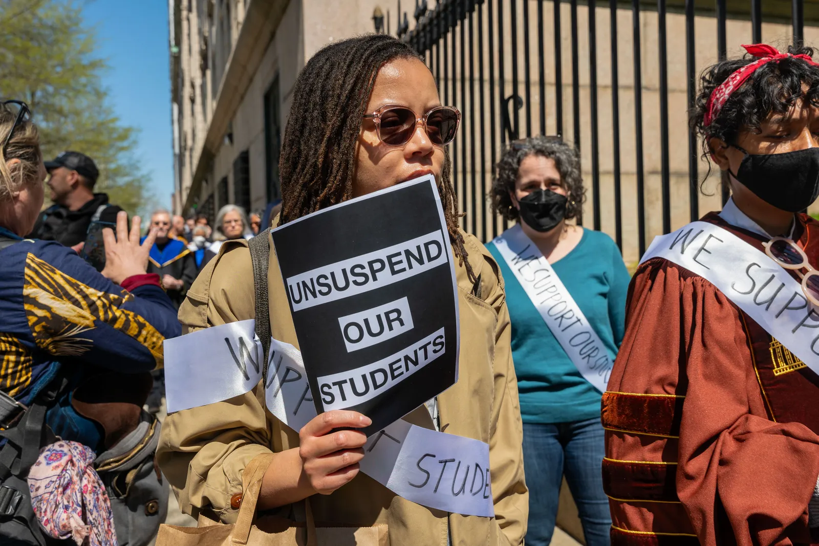 A demonstrator in a crowd holds a sign reading, “UNSUSPEND OUR STUDENTS.”