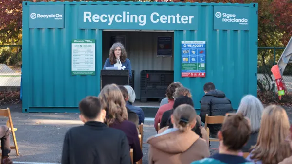 A woman speaks at a poduim in front of a shipping container converted into a recycling drop-off space.