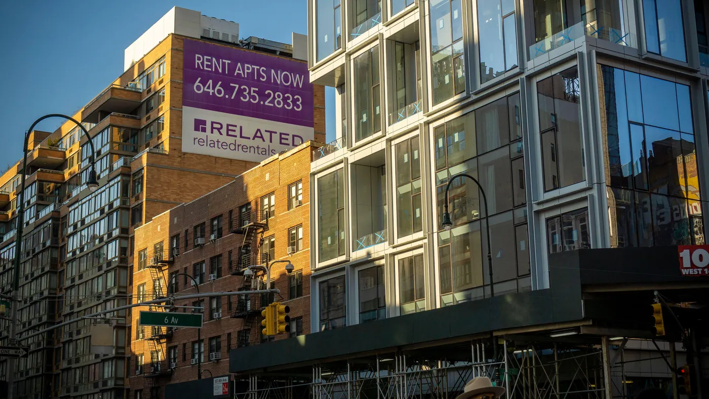Sunlight casts a shadow on brick and glass-front apartment buildings against a blue sky, with a 6th Avenue street sign in the foreground