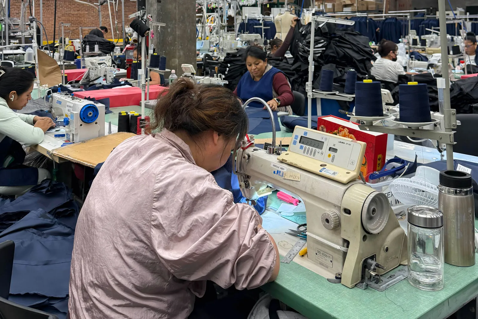 Workers use sewing machines to make blue clothing in a large factory filled with stacks of pre-cut fabric.