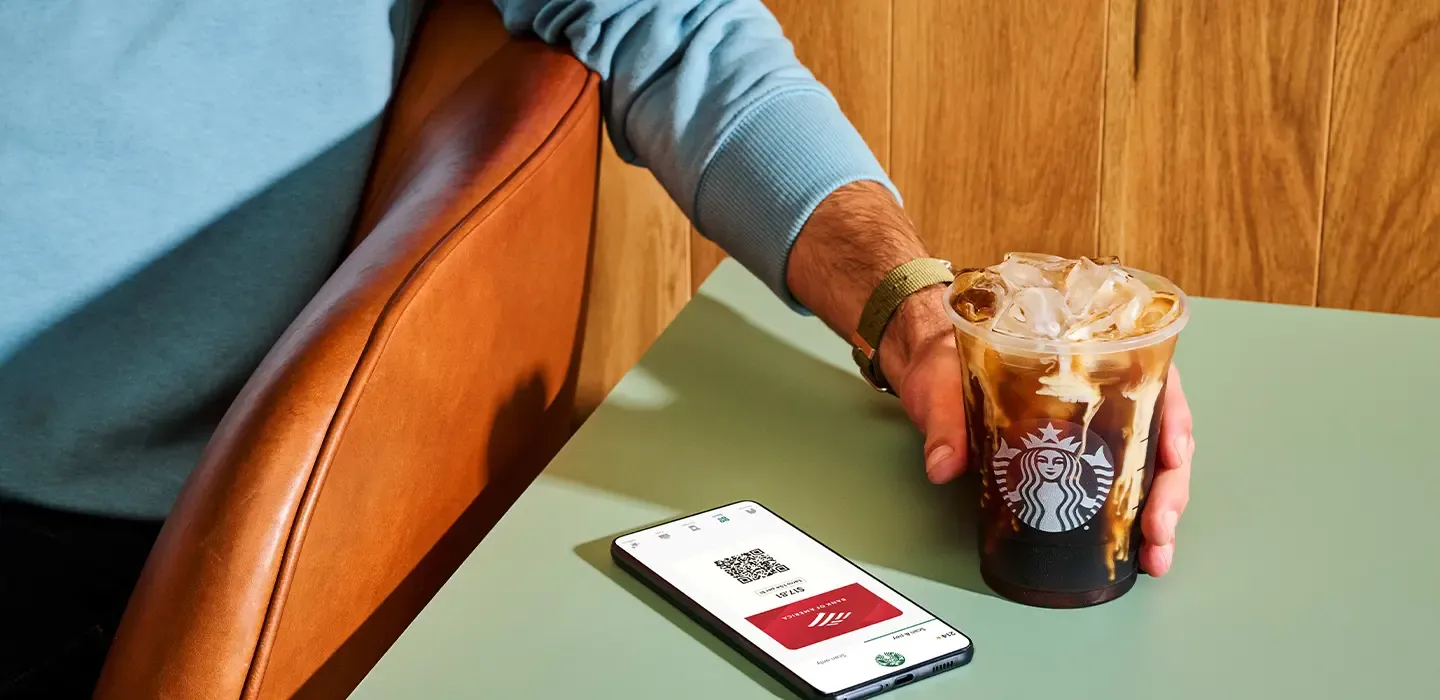 A man holds a starbucks drink next to a phone with a Bank of America card onscreen.