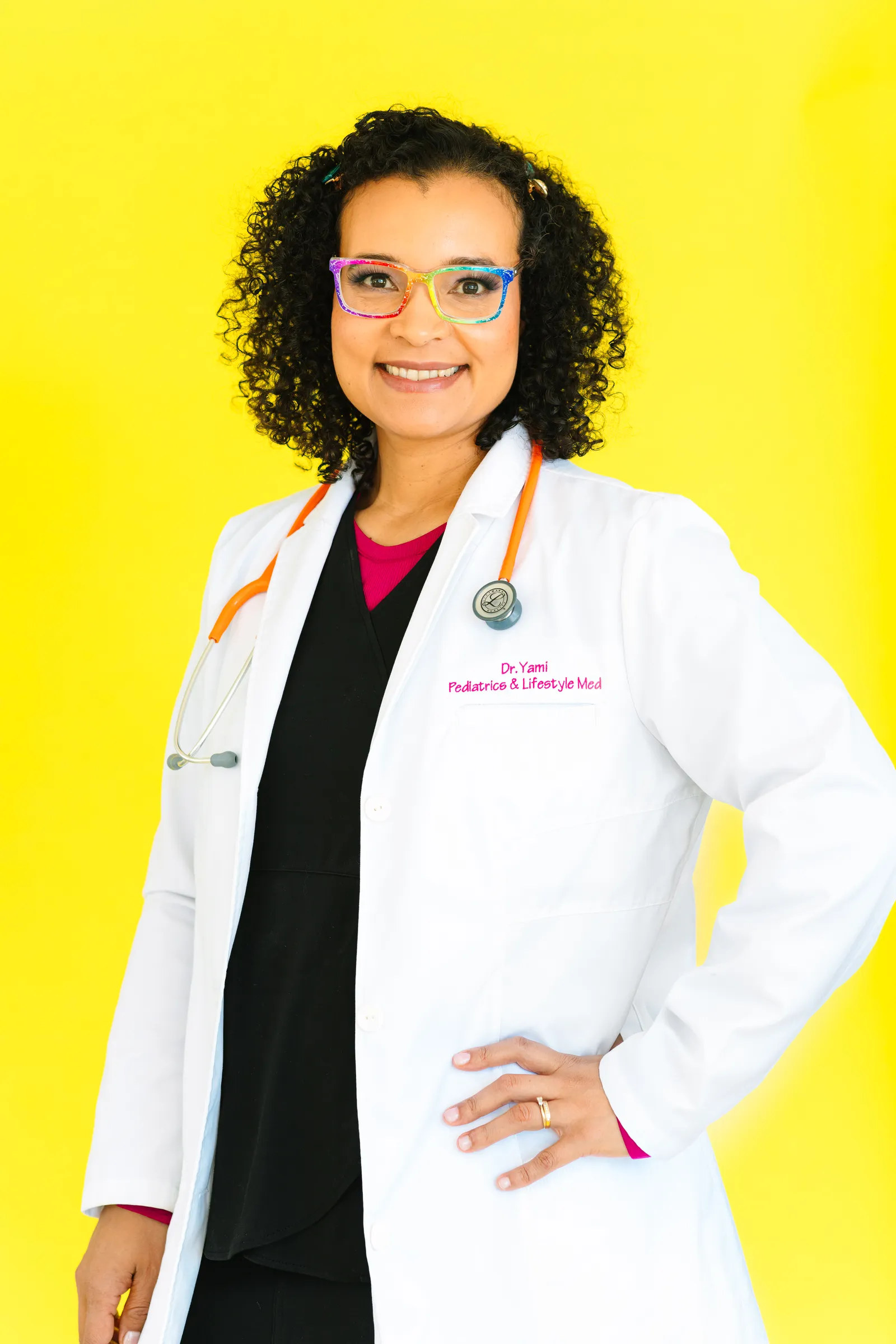 A woman pediatrician stands in front of a yellow background for a portrait photo.