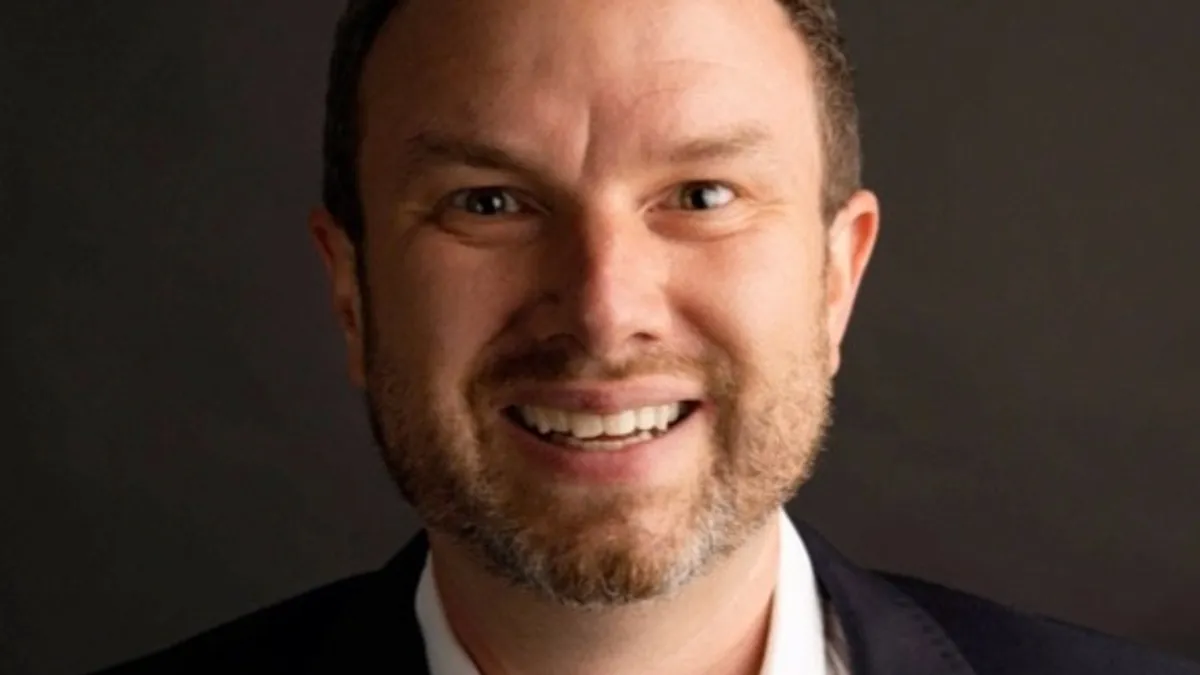 A man, who really needs collar stays or a tie, grinning and wearing a white shirt under a navy or black sportcoat.