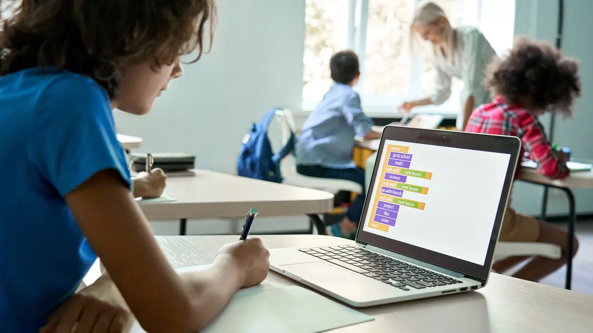 Student seated at desk in classroom looks at laptop computer screen and holds a writing instrument in the right hand. Other students and an adult are in the background.