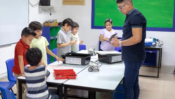 A handful of students are standing around a table in a classroom. An adult stands on the other side of the table. On the table are bins and a robot.