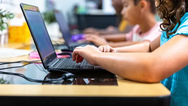 Students sit at a table in a row. They are working on laptops