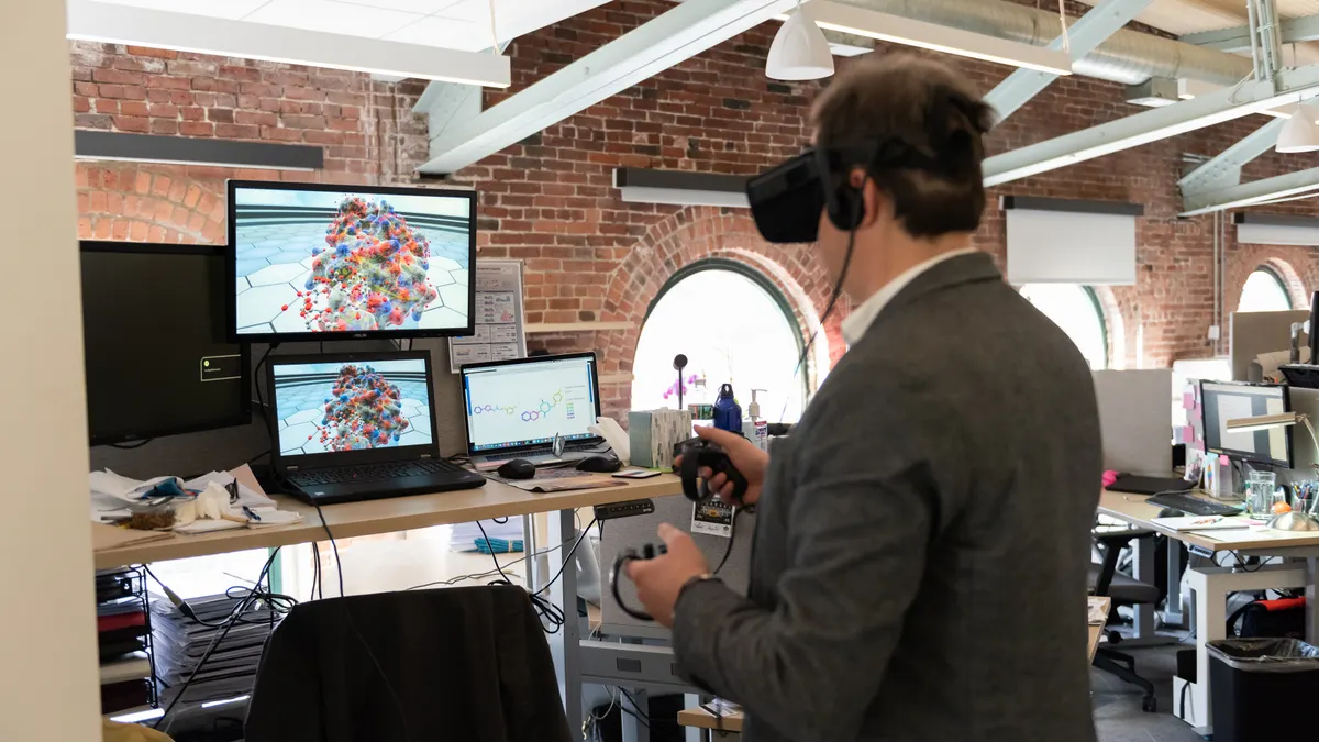 A person in an office uses a virtual reality headset to examine a 3D model on a computer screen.