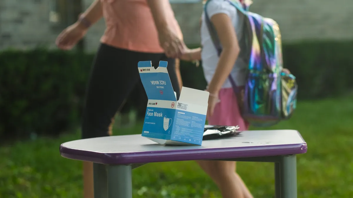 A box of face masks sit on a table outside. In the background, an adult and student appear to be walking by the table.