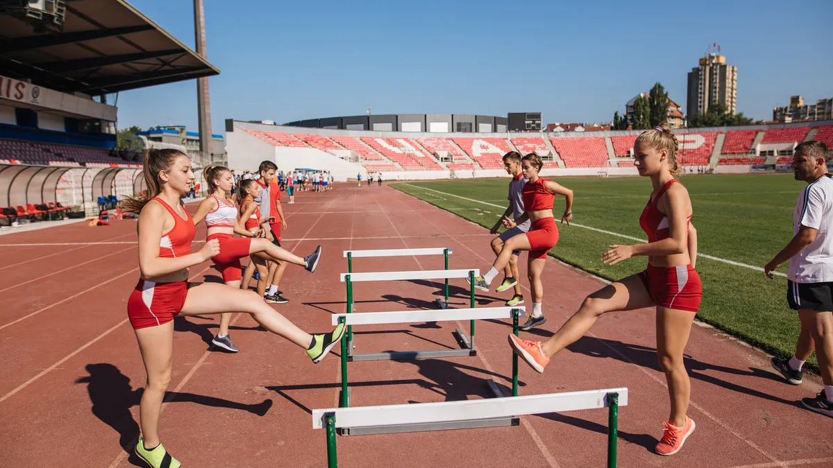 Young athletics team hurdling on a sports stadium, exercising together, overlooked by a sports coach.