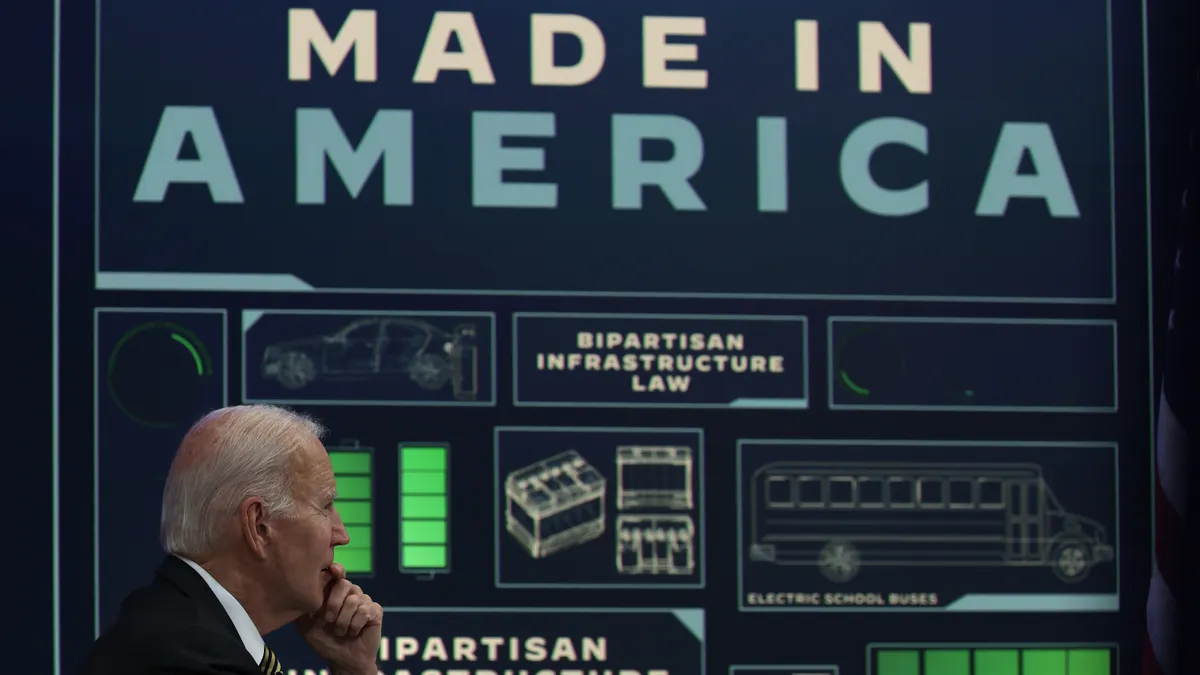 President Joe Biden sits at a desk. In the background, a sign can be seen reading "Made in America" with pictures of batteries and automobiles.