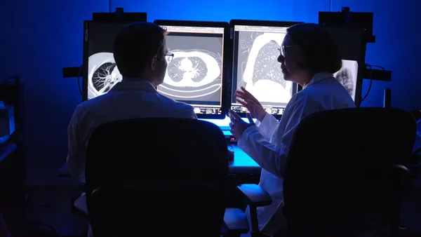 Two people face each other and speak in a dark room in front of screens showing a black and white image of a lung.