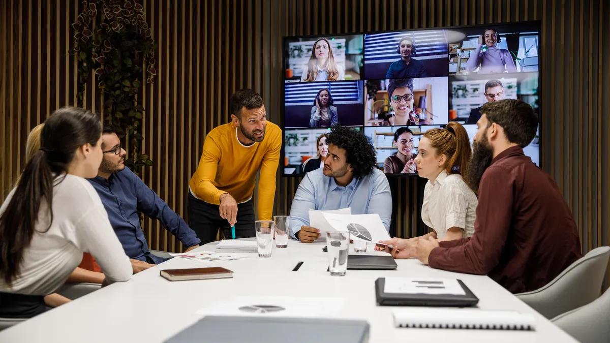 A group of employees discuss an upcoming project during a meeting with some colleagues attending via video conference.