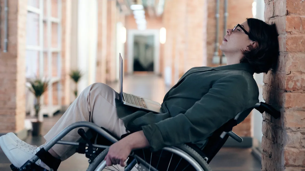 A person leans back in their wheelchair, with a laptop on their lap, looking stressed and tired