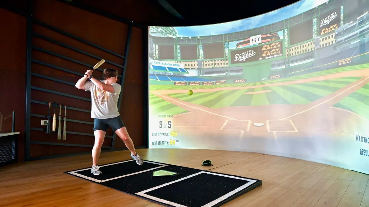 A person wearing a white shirt holds a bat while a green ball floats in the air. There is a projection screen on the right with a baseball field.